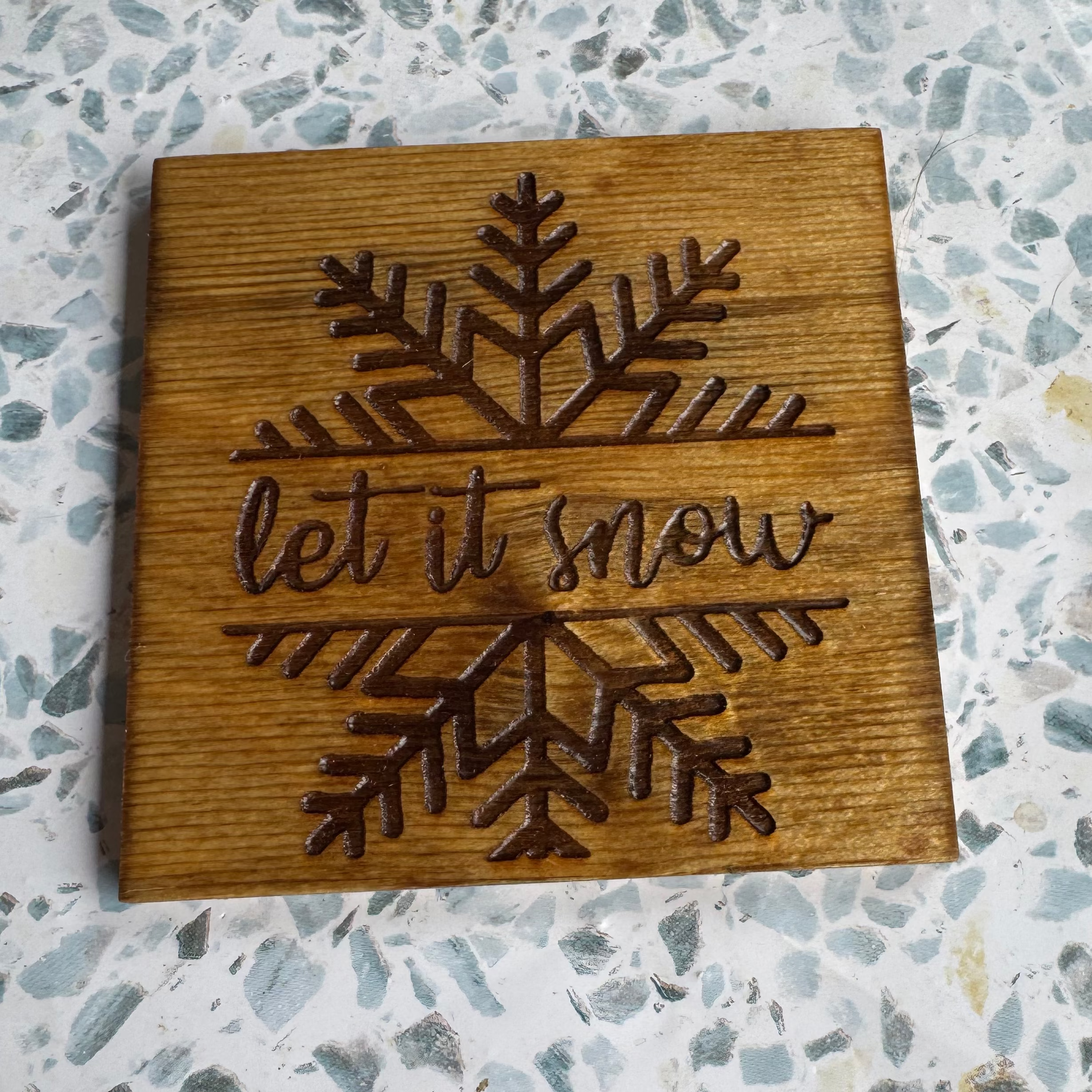 Wooden coaster with 'let it snow' and snowflake design on a textured surface